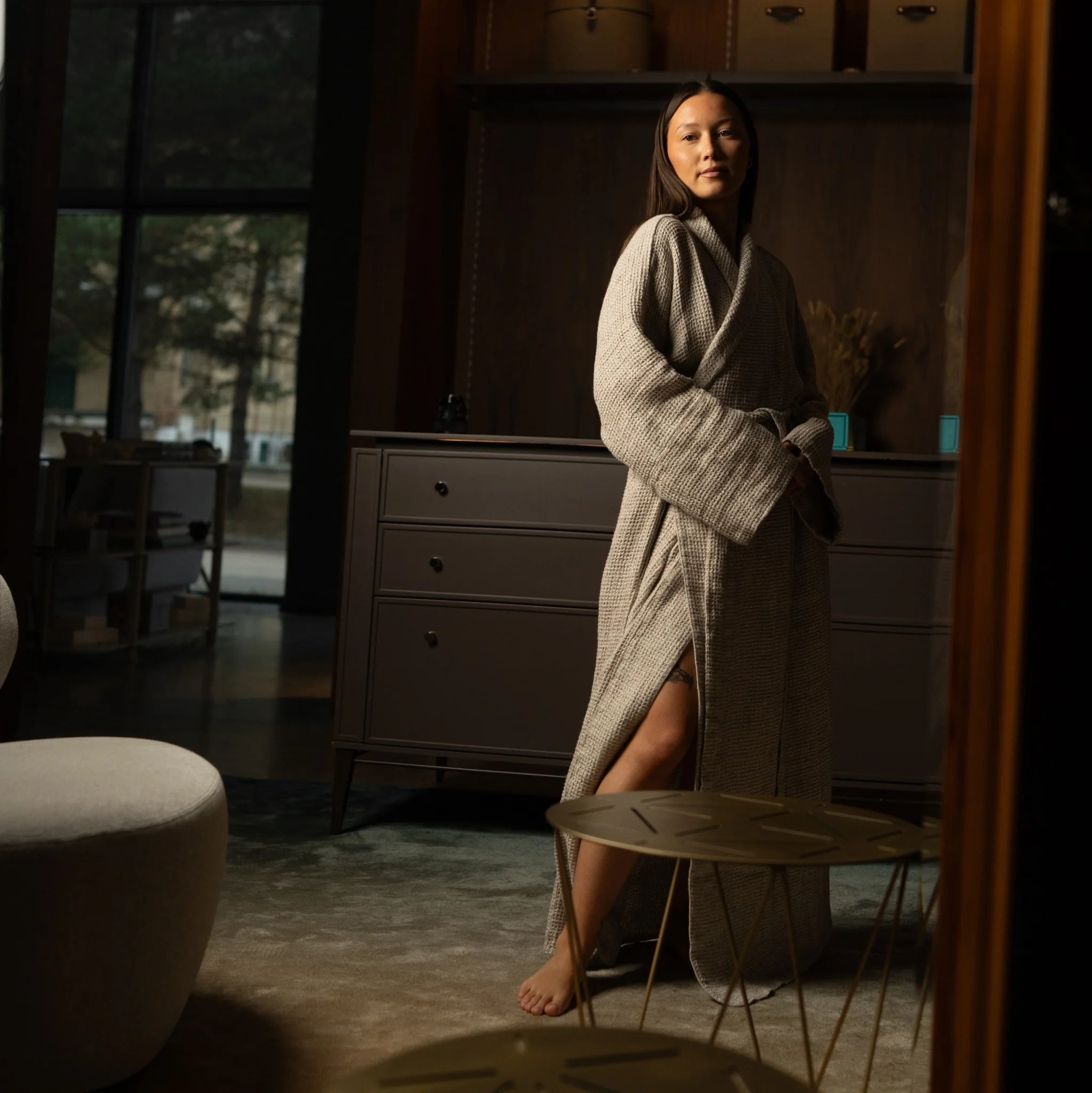 Woman standing in front of a mirror wearing a beige unisex linen robe in a calm, minimal interior